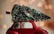 A red toy car carries a Christmas tree, set against a warm bokeh background, celebrating the festive season.